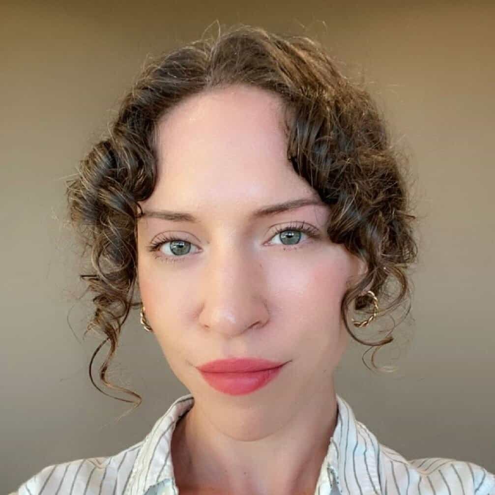 A person with curly brown hair, wearing gold hoop earrings and a white striped shirt, looks directly at the camera against a plain beige background.