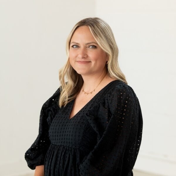 A woman with long blonde hair wearing a black dress with textured fabric stands in front of a plain white background, smiling softly at the camera.