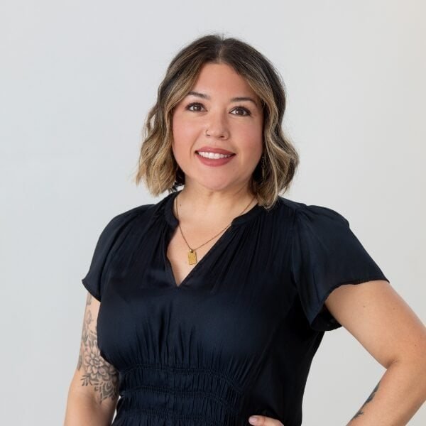 Smiling woman with short, wavy brown hair wearing a black blouse and gold necklace, standing with one hand on her hip against a plain light background. Tattoos are visible on her arms.
