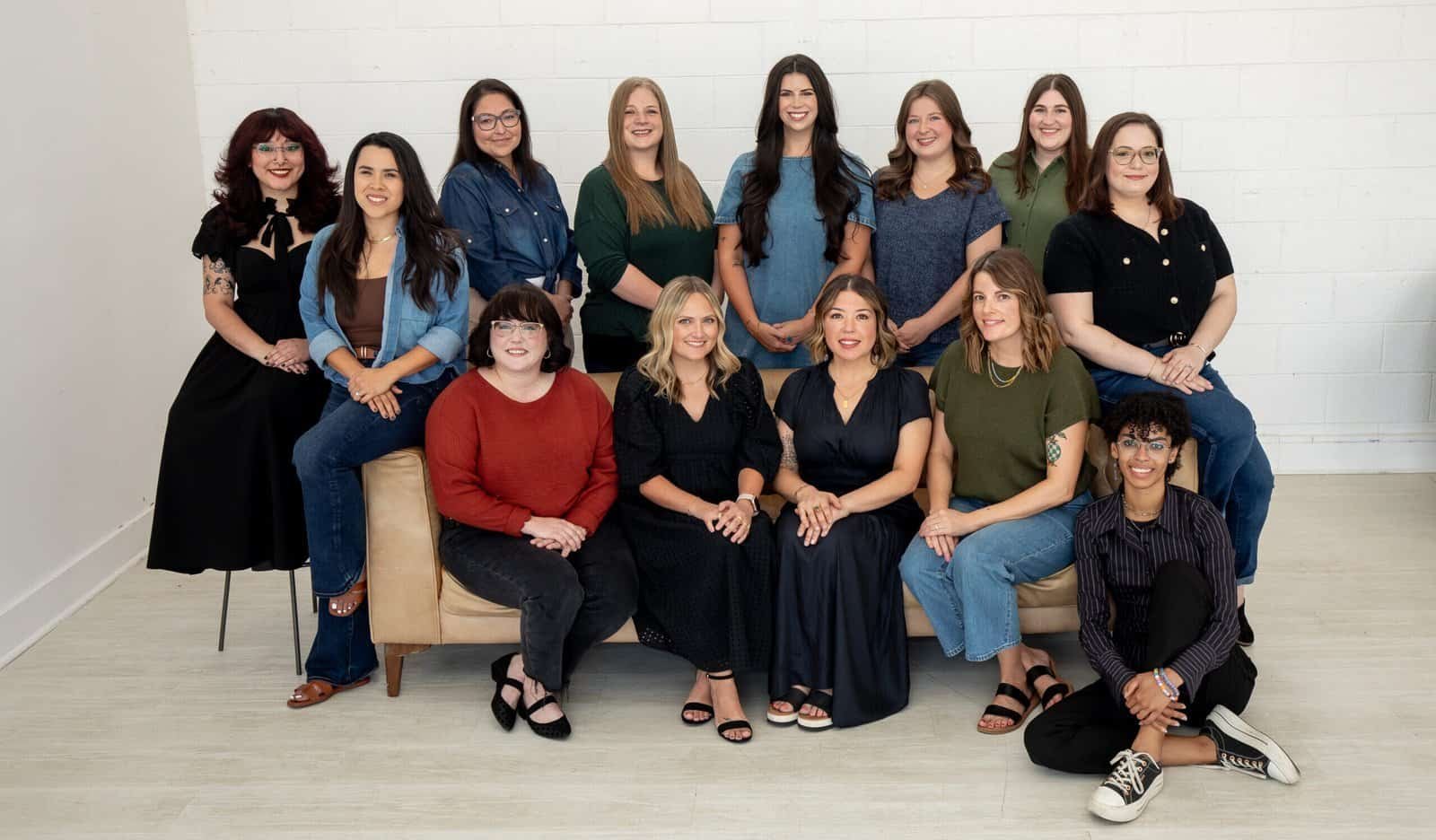 A group of 13 women pose for a portrait in a bright room with a white wall. Some are seated on a sofa and the floor, while others stand behind them, all smiling at the camera.