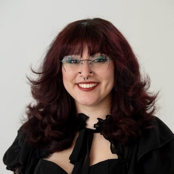 A person with wavy, dark red hair and bangs smiles at the camera. They wear glasses, have a septum piercing, wear dark lipstick, and are dressed in a black outfit with a bow at the collar against a plain background.