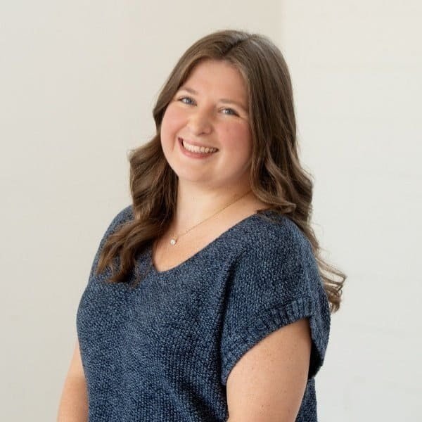 A woman with long brown hair is smiling at the camera. She is wearing a short-sleeved, blue knit top and a necklace, standing against a plain, light-colored background.