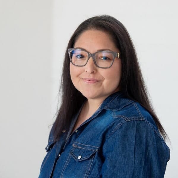 A woman with long dark hair and glasses is smiling slightly. She is wearing a denim jacket and standing in front of a plain light-colored background.