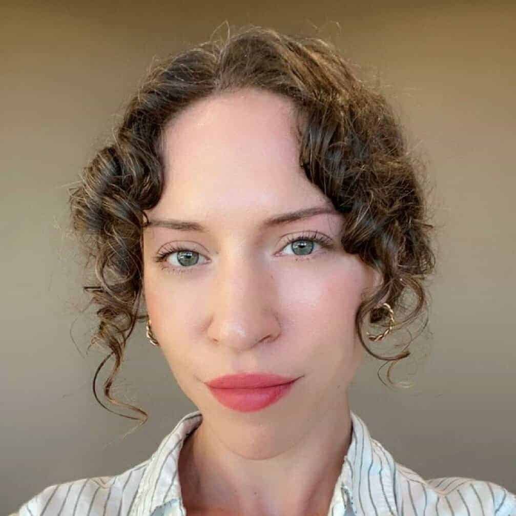 A person with curly brown hair, light eyes, and pink lipstick looks directly at the camera, wearing a light-colored striped shirt and gold hoop earrings, with a neutral beige background.