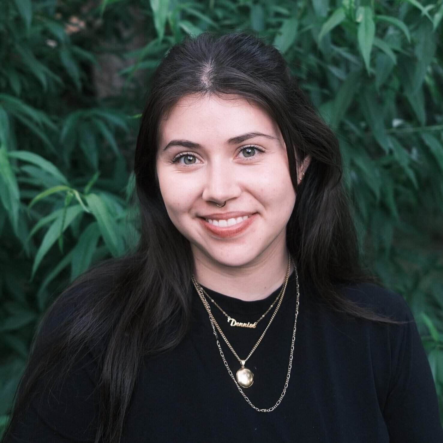 A woman with long dark hair, wearing a black top and layered necklaces, smiles in front of leafy green plants.