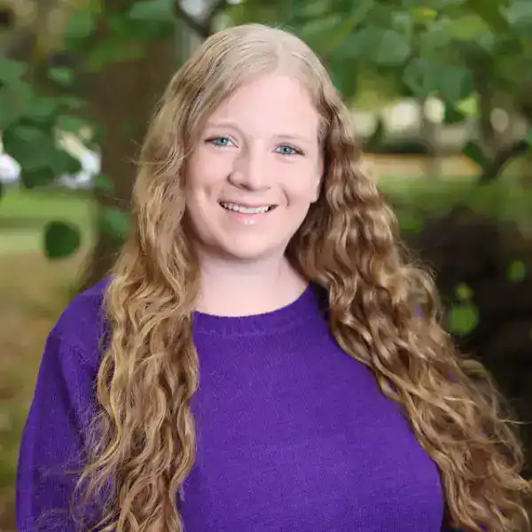 A smiling person with long curly hair wearing a purple sweater stands outdoors in front of green trees and foliage.