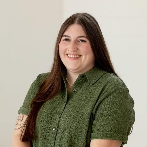 A woman with long brown hair, wearing a textured green shirt, smiles at the camera against a plain light background. She has a visible tattoo on her left arm and a small nose ring.