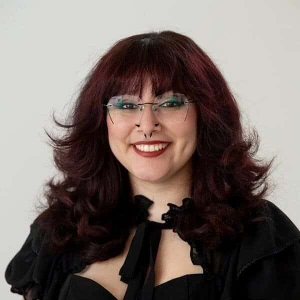 Smiling person with wavy, dark red hair, glasses, and a black outfit, standing against a plain white background.