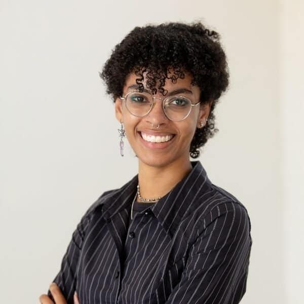 A person with short curly hair, glasses, and earrings smiles with arms crossed, wearing a dark pinstripe shirt against a plain light background.