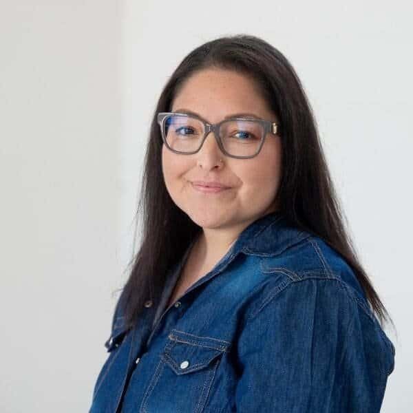 A woman with long dark hair and glasses smiles softly. She is wearing a blue denim jacket and is positioned against a plain white background.