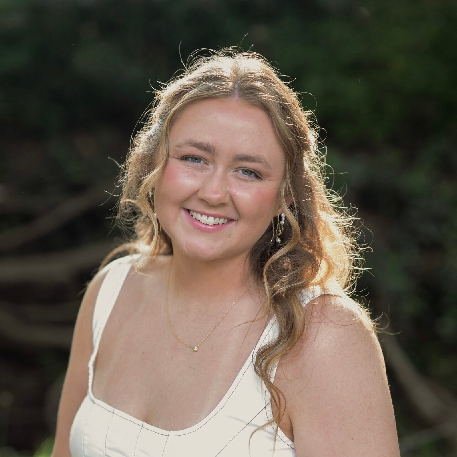 A smiling young woman in a white tank top poses for a portrait outdoors.